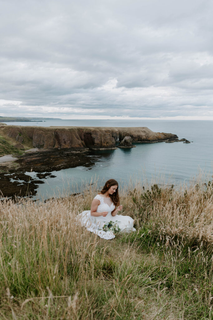 A woman sitting in her wedding dress on the cliffs along the east coast of Scotland during her Scottish elopement