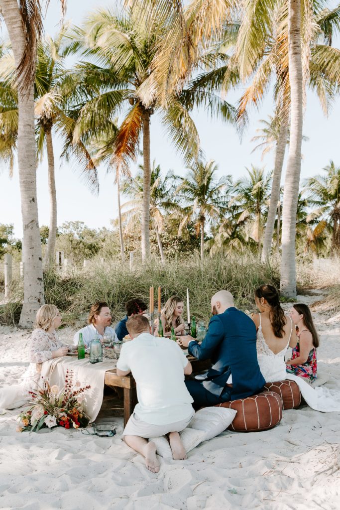 a newly wed couple and their friends enjoying an ocean side picnic after their beach ceremony