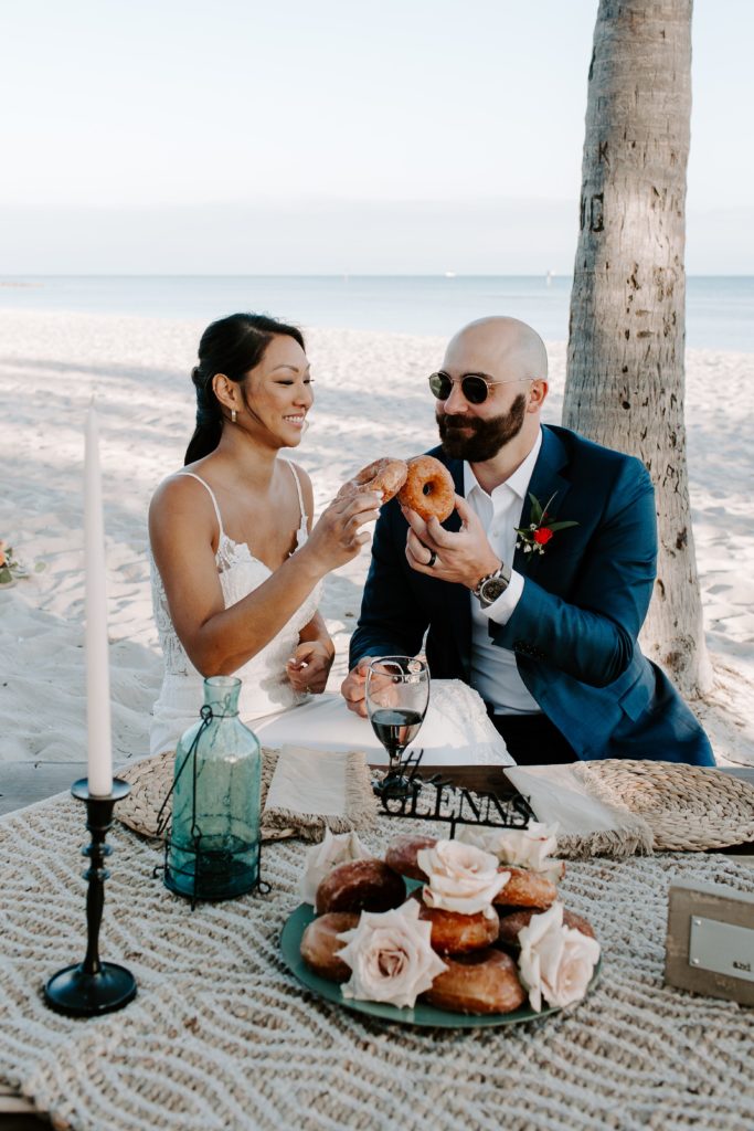 Couple cheering doughnuts after as they sit down for a beach picnic after their Florida keys elopement