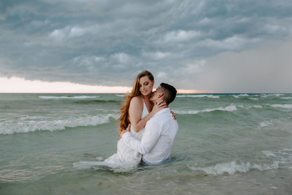 Woman sitting on top of her partner as he kisses her neck both of them sitting in the ocean with a storm rolling into the sky during their sunset summer elopement