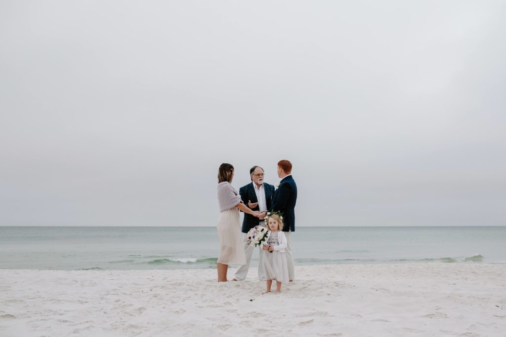 Couple holding their hands during their ceremony on 30A beach while their daughter holds her moms bouquet