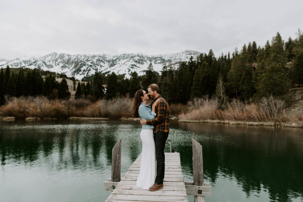 couple nuzzling their noses together as they stand on a dock overlooking a mountain range and pine trees during their Montana all day elopement