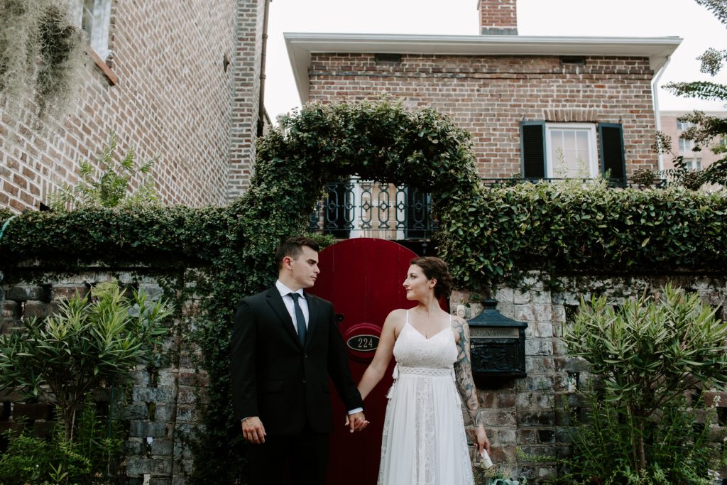 couple holding each others hands while they are looking at each other while standing in front of a red door during their all day urban elopement in Savannah, Georgia