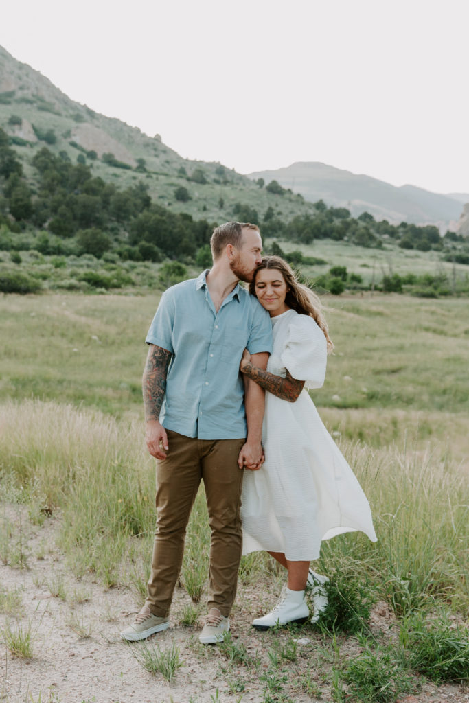 A man and a woman standing in a grass field and the woman is holding onto her partners hand as the man gives her a kiss on the temple during their Colorado Springs couple photos at Garden of the Gods