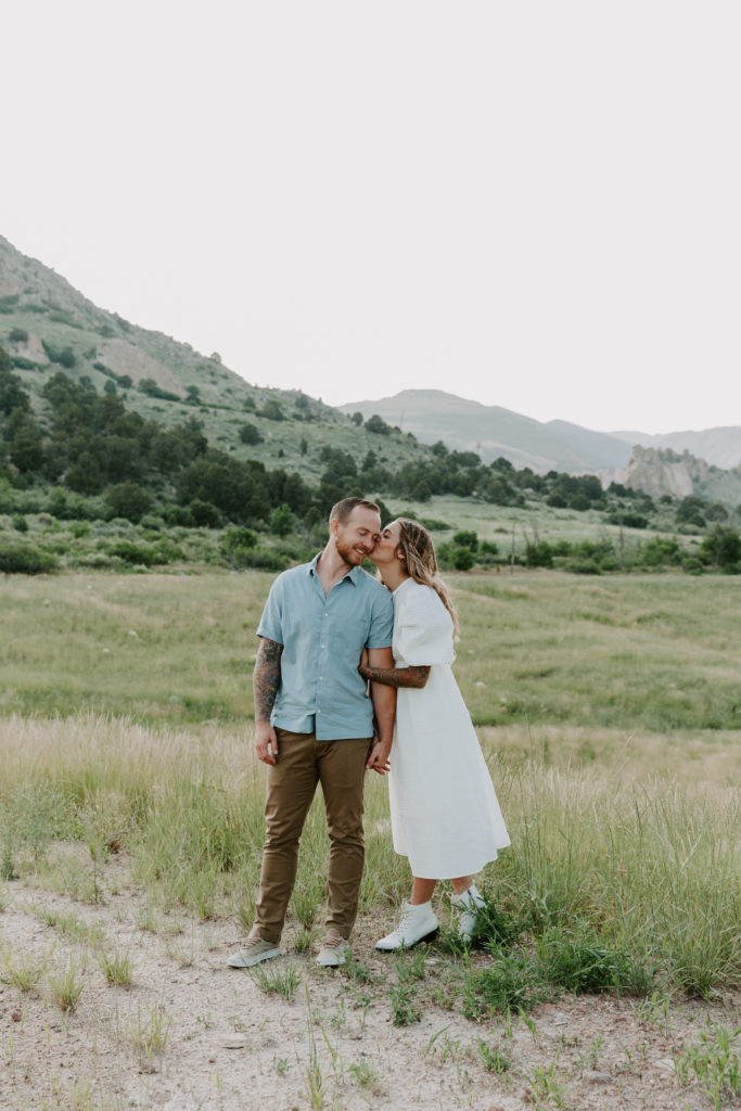 Couple standing in a field while they hold hands and the woman gives her partner a kiss on the check during their Colorado adventure session