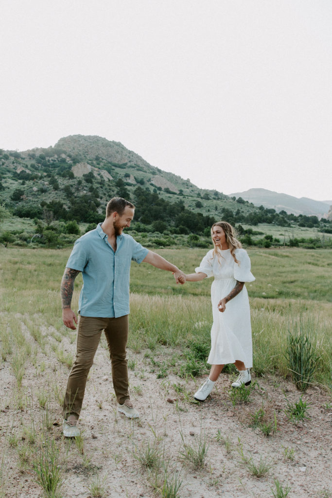 Couple holding hands and walking as they are laughing at each other with the mountains in the background during their midwest engagement photos