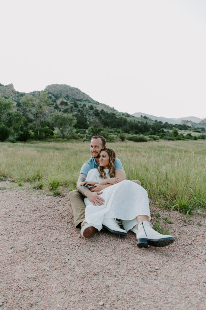 Couple sitting on a dirt path cuddling with the woman sitting between his legs while her partner has his arms wrapped around her and they looking off into the distance during their Colorado engagement photos