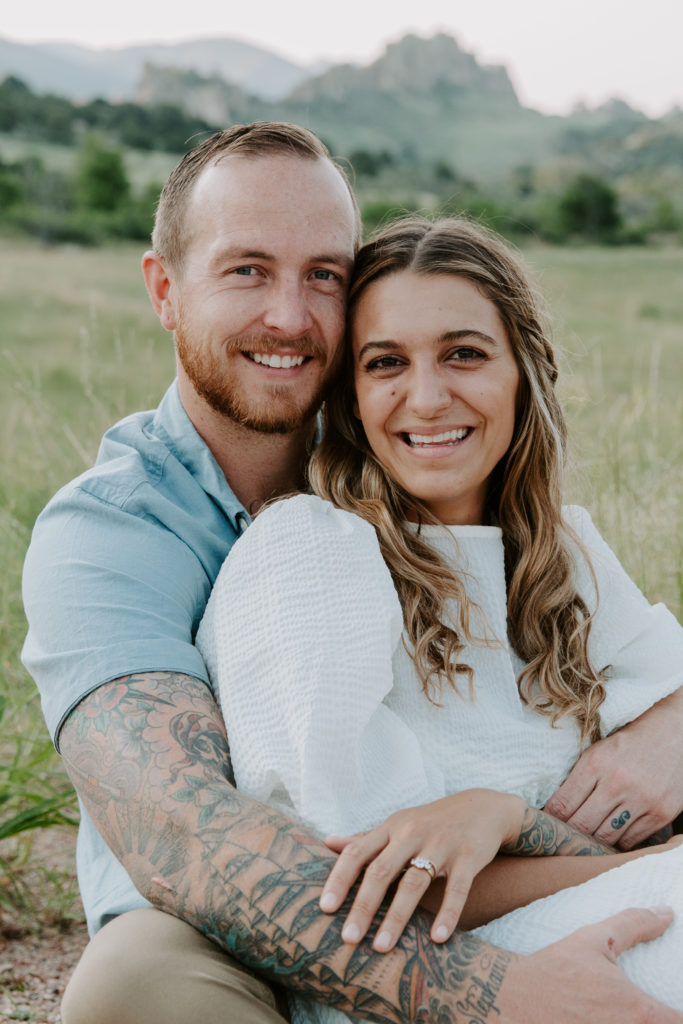 Couple sitting on a dirt path with the woman sitting between his legs while her partner has his arms wrapped around her and they are smiling during their Colorado couple photos