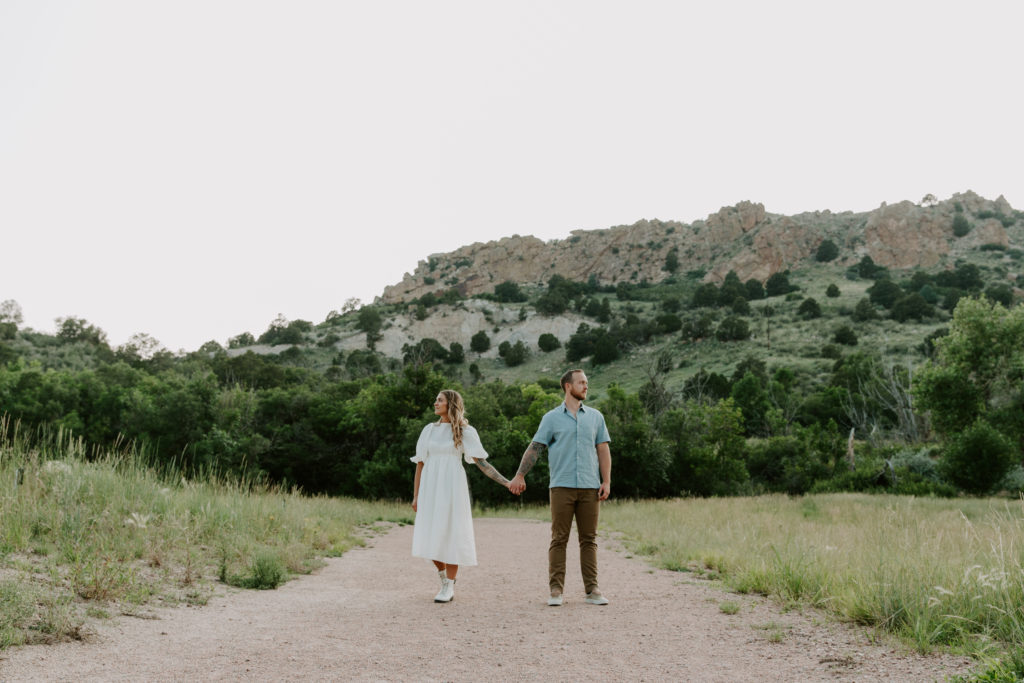 Couple standing on a dirt path while holding hands and looking the opposite directions of each other with the Colorado mountains in the background