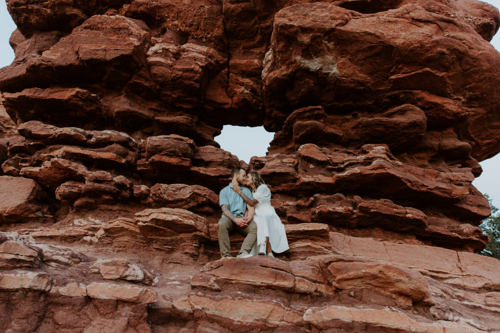 Couple sitting on the edge of red rock formations with the woman caressing her partners face during their Colorado engagement photos