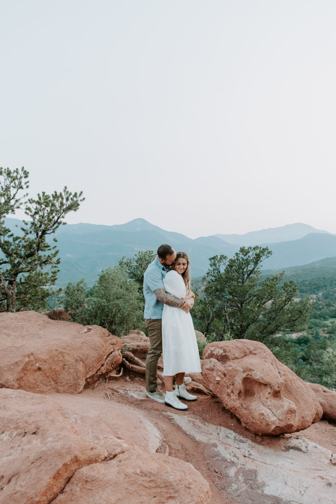 A man holding his partner from behind as she curls up into him with the red rocks beneath them and the mountains in the background during their Colorado engagement photos