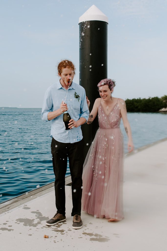 Couple popping champagne on a dock as the man trys to spray it in his mouth and his new wife is laughing