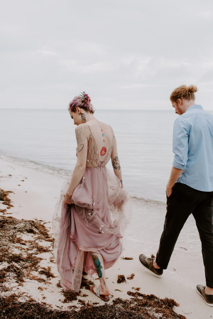 Couple in wedding attire walking along the edge of the beach looking for seashells