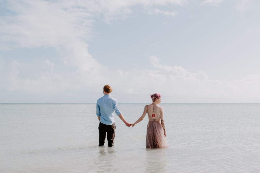 Couple standing in the ocean with nothing but their hands touching while they look opposite directions of each other