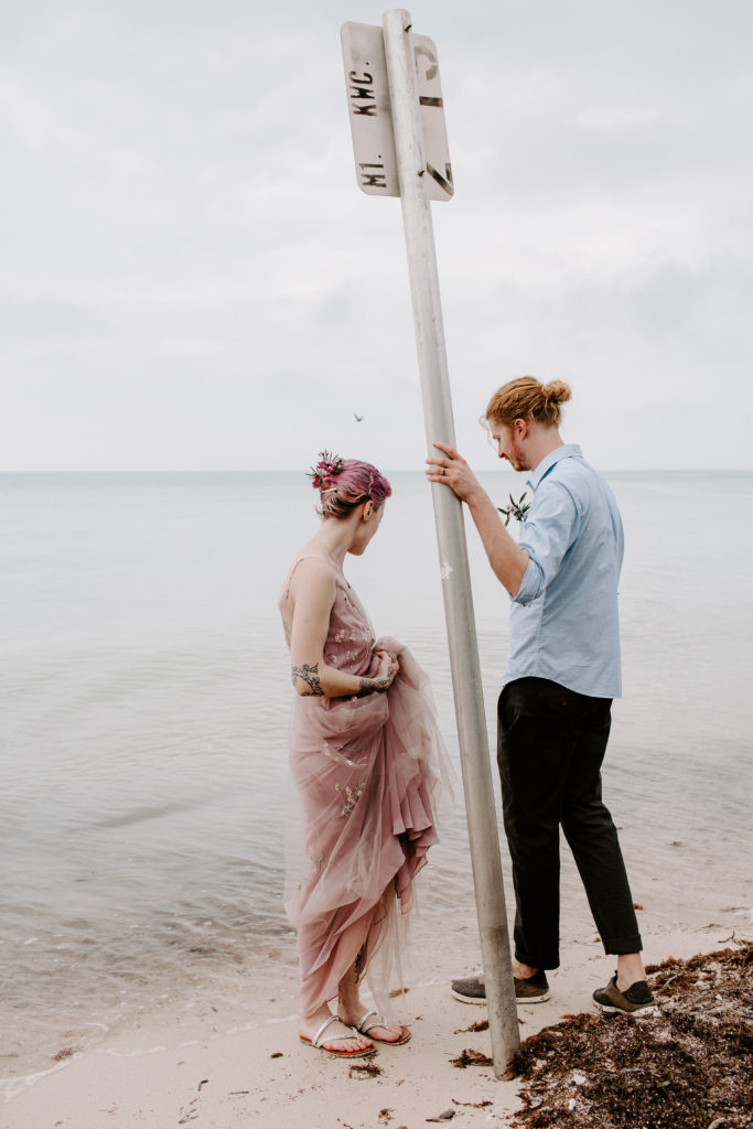 Man holding on to a pole that is randomly on the beach while the woman holds her dress up from getting wet during their Friday the 13 key west elopement