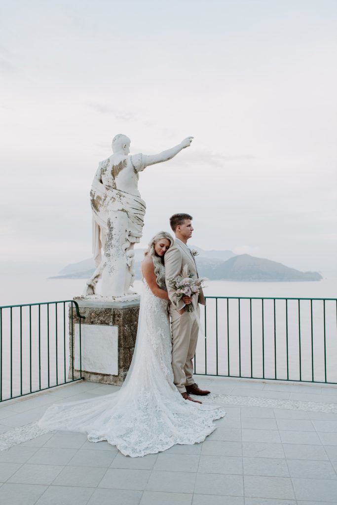 Woman hugging onto the back of her partner with the sea behind them and the statue next to them after they got married on the Amalfi coast