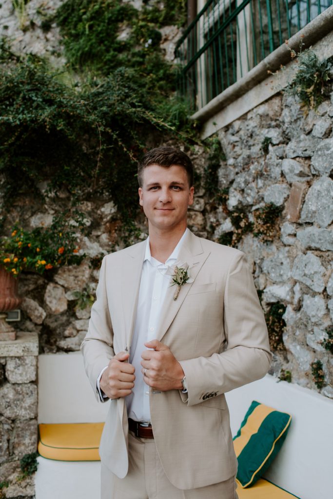 Man holding the sides of his tan suit jacket soft smiling with a rocky background after he got married on the Amalfi coast