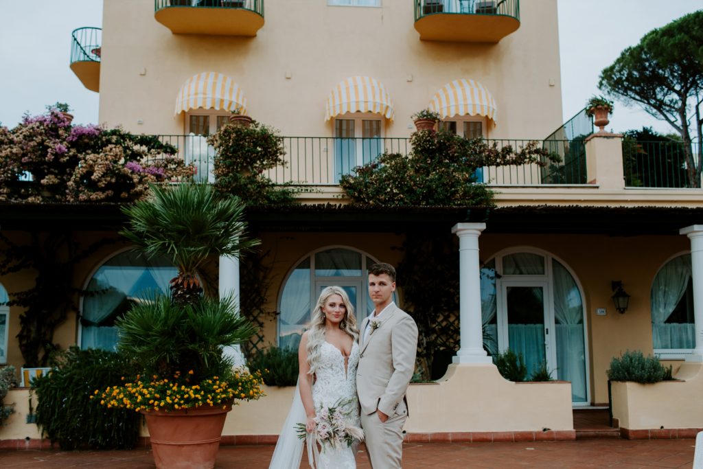 Bride and groom standing with their arms around each other looking like the cover of Vogue standing in front of a yellow building during their European elopement