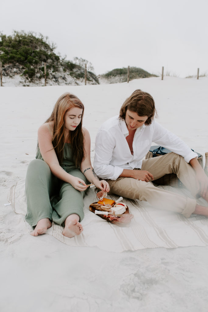 A man and a woman making a snack from the charcuterie board beach picnic during their beach photoshoot