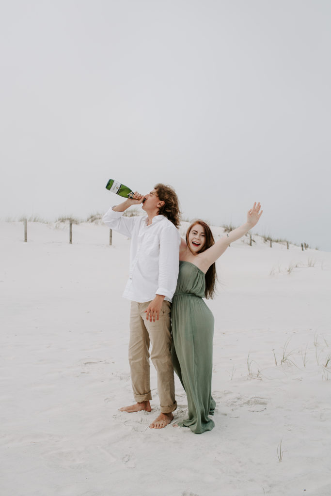 A woman cheering on her partner as he takes a big drink of a bottle of champagne during their Destin couple photos