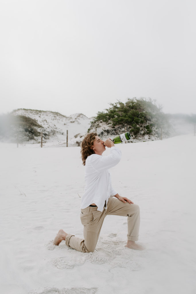 A man on one knee chugging a bottle of champagne during his beach photoshoot in Destin, Florida