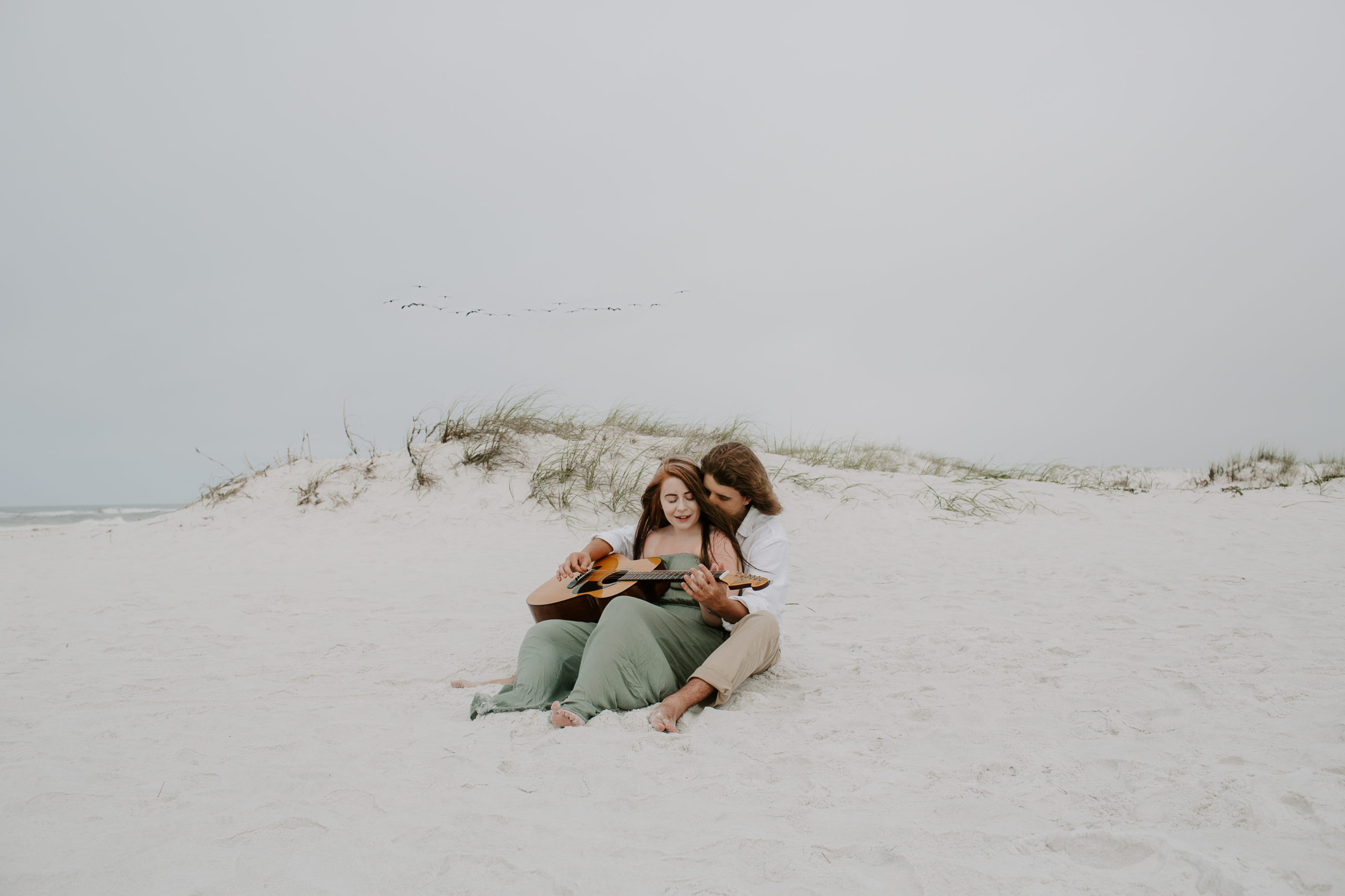 A couple sitting in the sand with the large dunes in the background playing the guitar