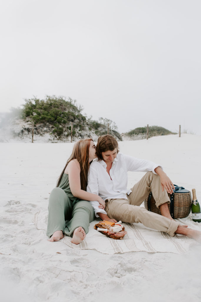 A woman giving her partner a kiss on the cheek as he is looking down and her arms are on his during their beach photoshoot in the Florida panhandle
