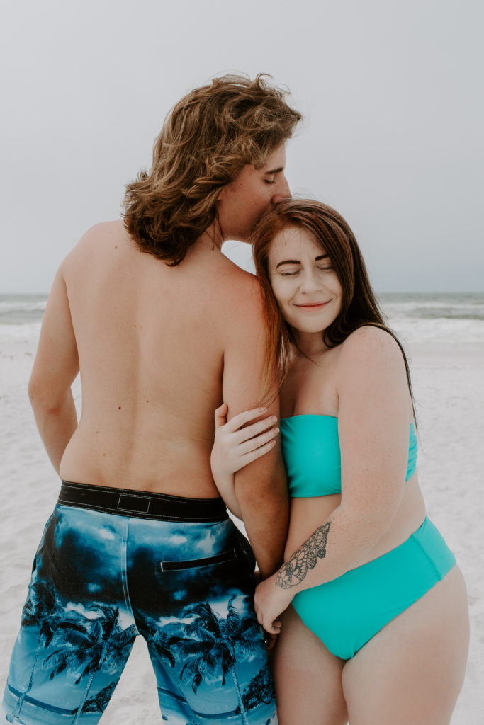 A woman holding onto her partners arm as he gives her a kiss on the head during their early morning swimsuit photoshoot
