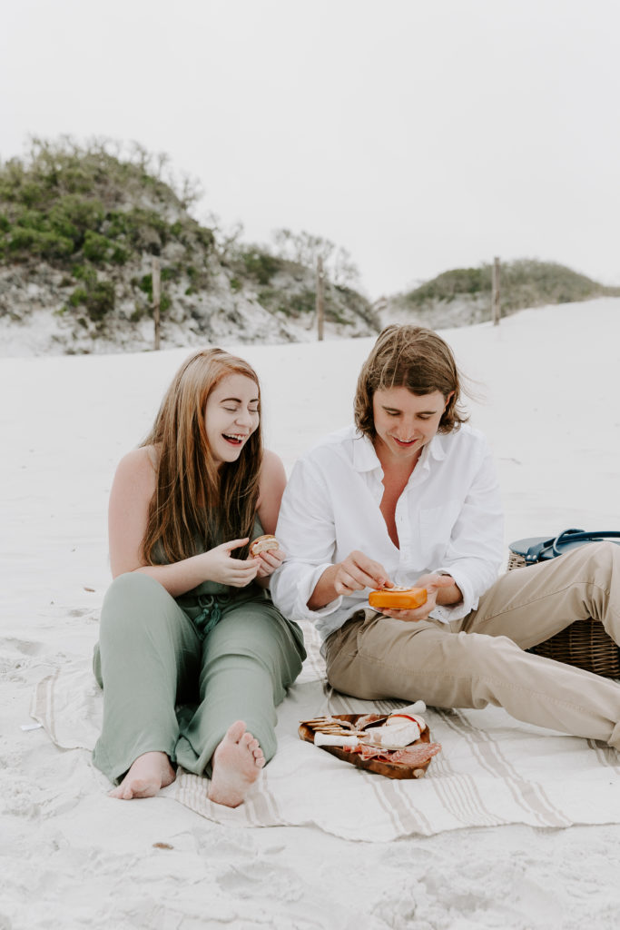 A woman laughing as her parter makes her a bite of food during their beach picnic photoshoot in Destin, Florida