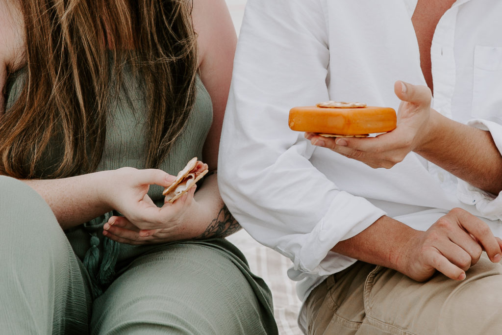 A woman holding a cracker and her partner holding a whole block of cheese during their beach picnic in Destin