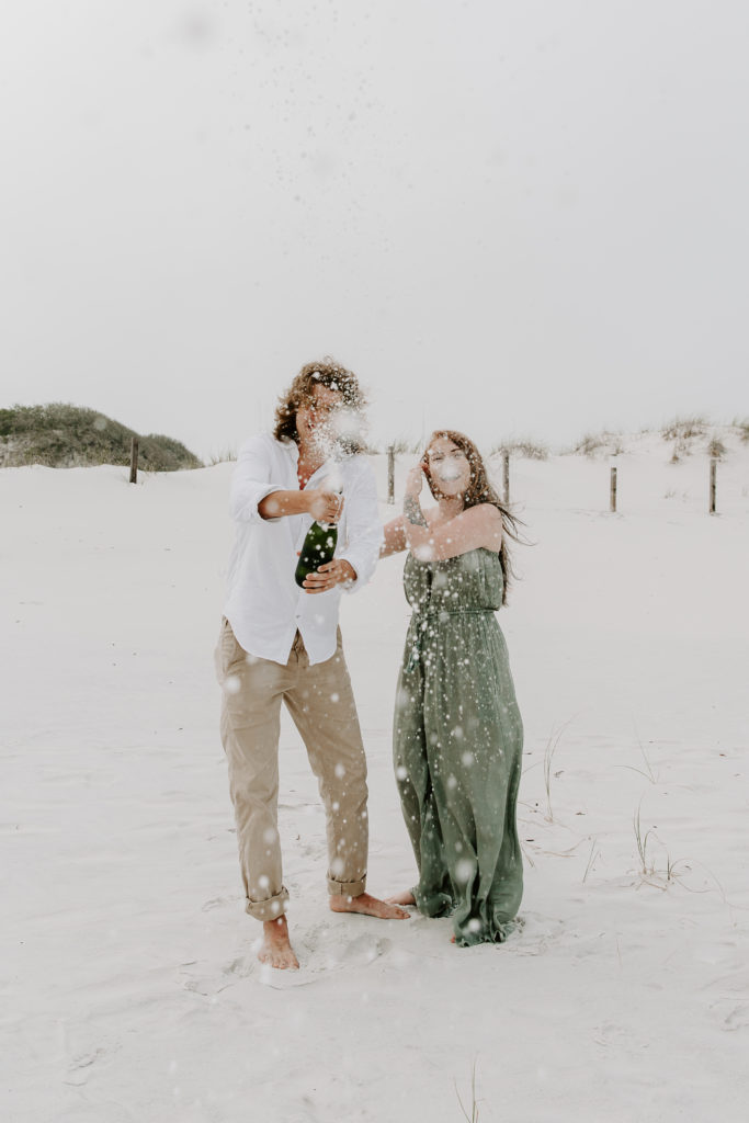 A man popping a bottle of champagne as the woman is pushing her hair behind her ear during their beach engagement photos in Florida