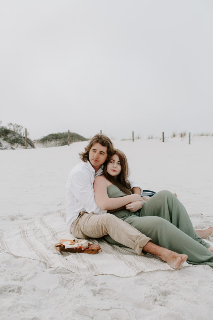 A couple sitting on the beach with the woman sitting on her partners lap as they both look off into the distance during their Okaloosa Island engagement photos