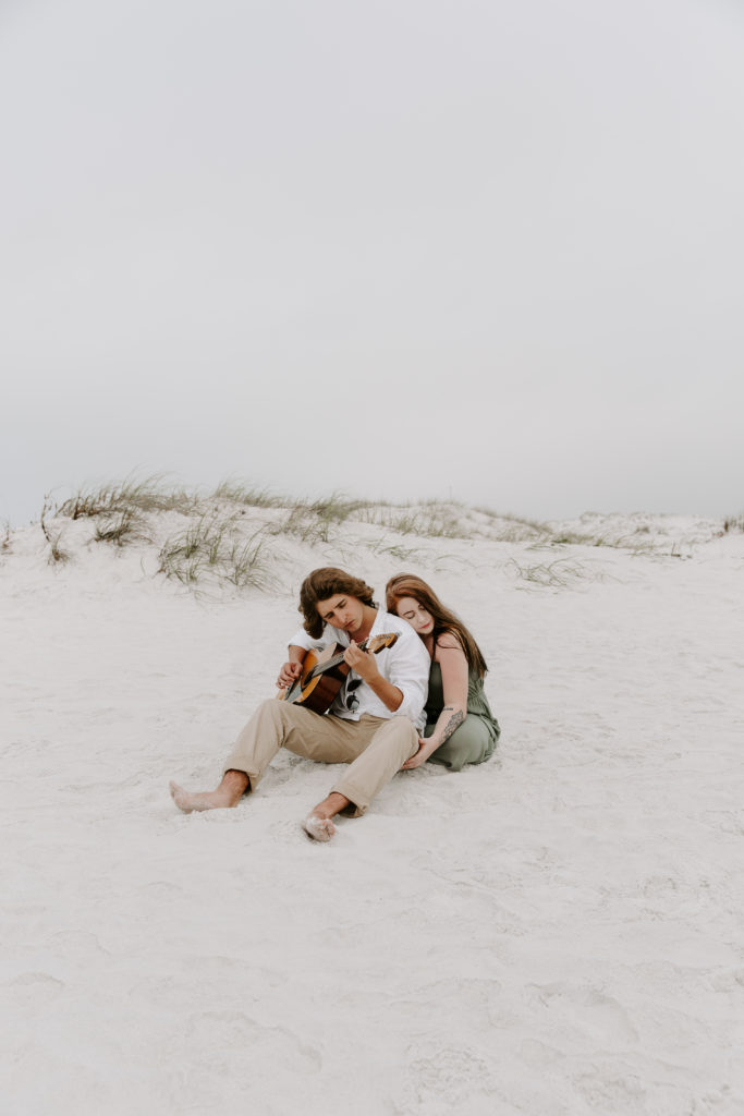 A woman leaning against her partner as he plays the guitar with the sand dunes in the background during their Destin engagement photos