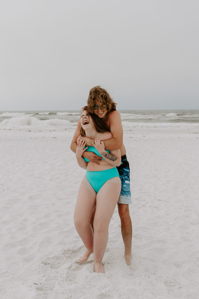 Woman laughing as her partner is hugging her from behind with the ocean in the background during their early morning beach photos in Destin, Florida