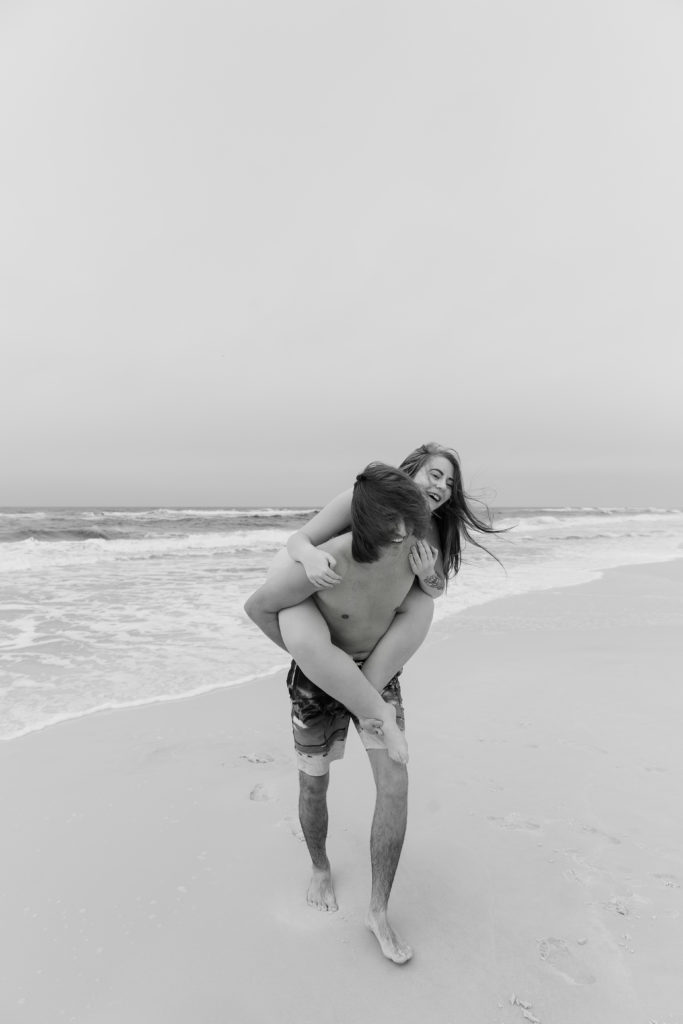 Man giving a woman a piggy back ride as they are both laughing and running away from the ocean during their beach photoshoot
