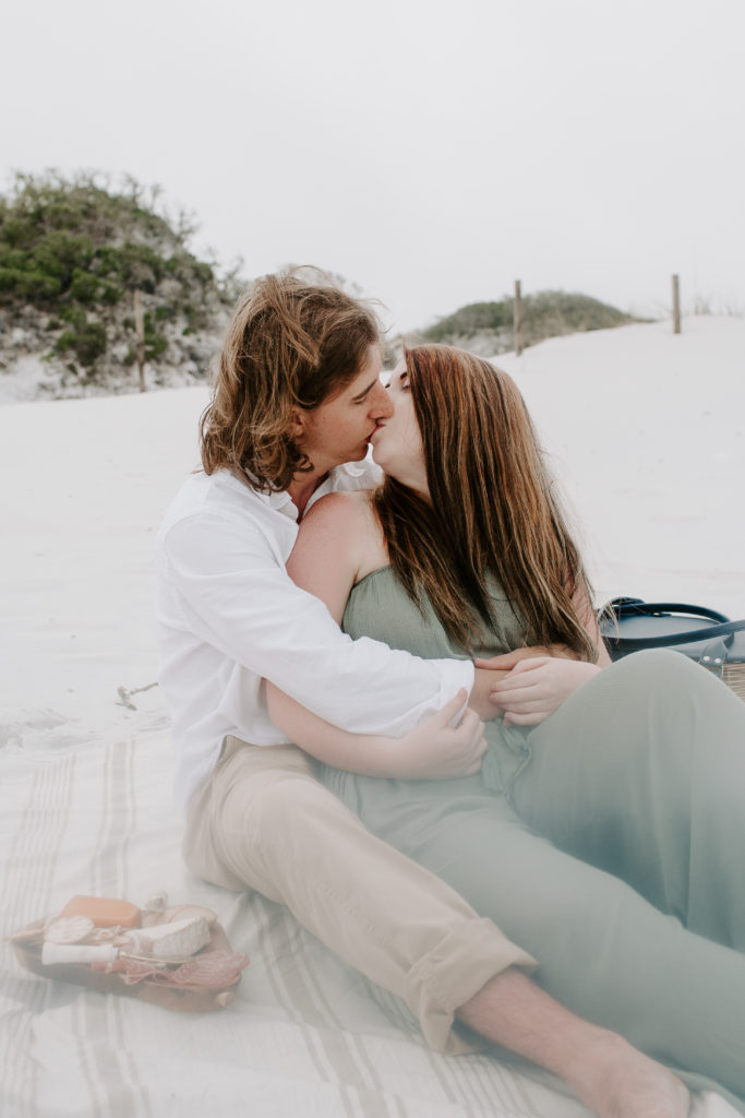 A couple sitting in the sand with their arms around each other sharing a kiss during their okaloosa island beach photoshoot
