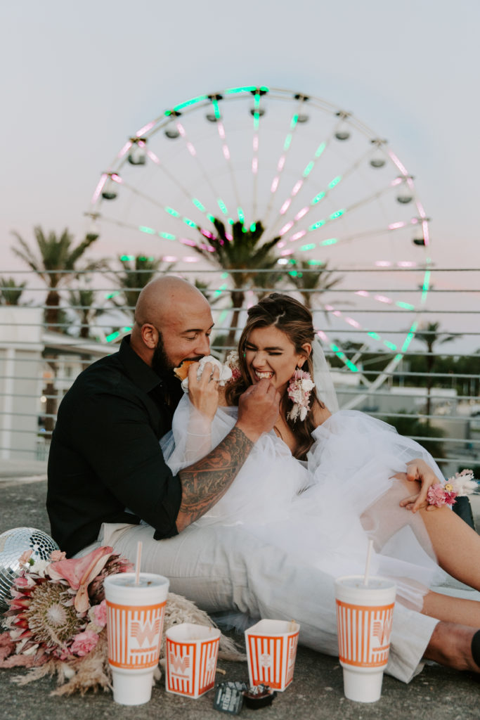 Man feeding his partner french fries as she is feeding him a burger with a ferris wheel behind them during their Alabama elopement
