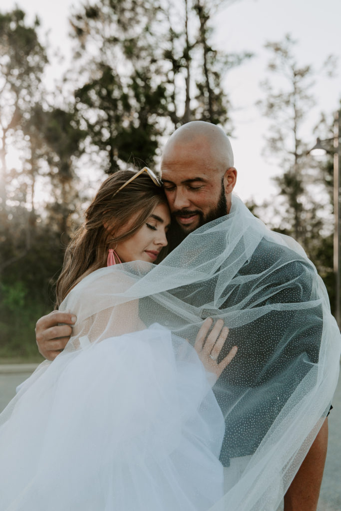 Couple wrapped up in a wedding veil with their eyes closed with the forest behind them after saying their vows