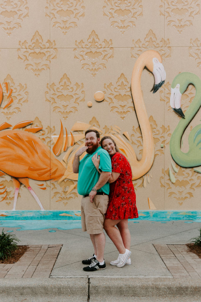 A woman cuddling up to her partners back as he is being silly and gasping with a flamingo mural in the background during their Destin engagement photos