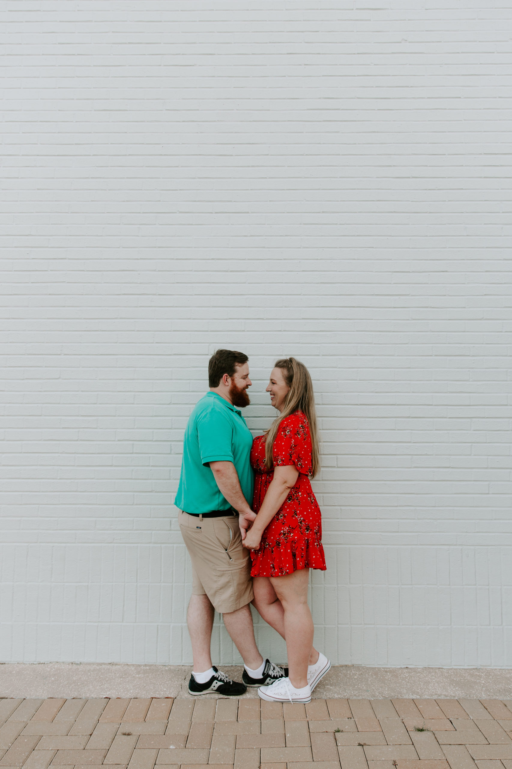 A couple leaning up against a white wall looking at each other and holding their hands during their engagement photos at the Destin Commons