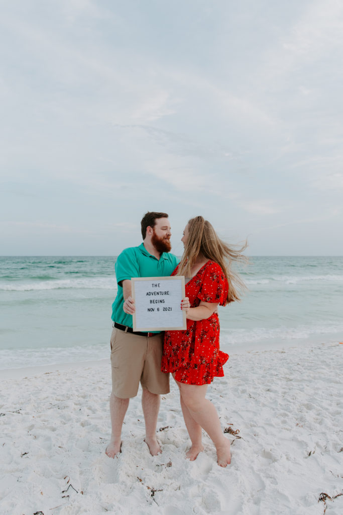 A couple standing on the beach holding out a sign that has their wedding date on it during their beach engagement photos