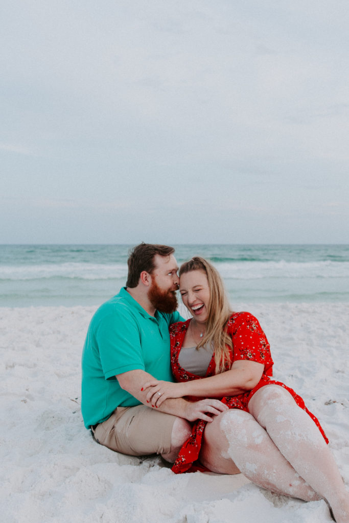 A woman sitting in her partners lap laughing hard as he is making faces and saying things into her ear during their sunset beach photos in Destin
