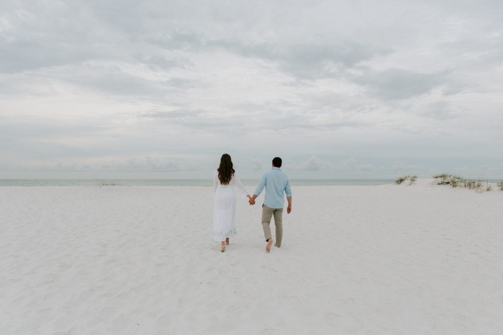 A newly engaged couple holding hands and walking off towards the horizon during their early morning beach couple photos