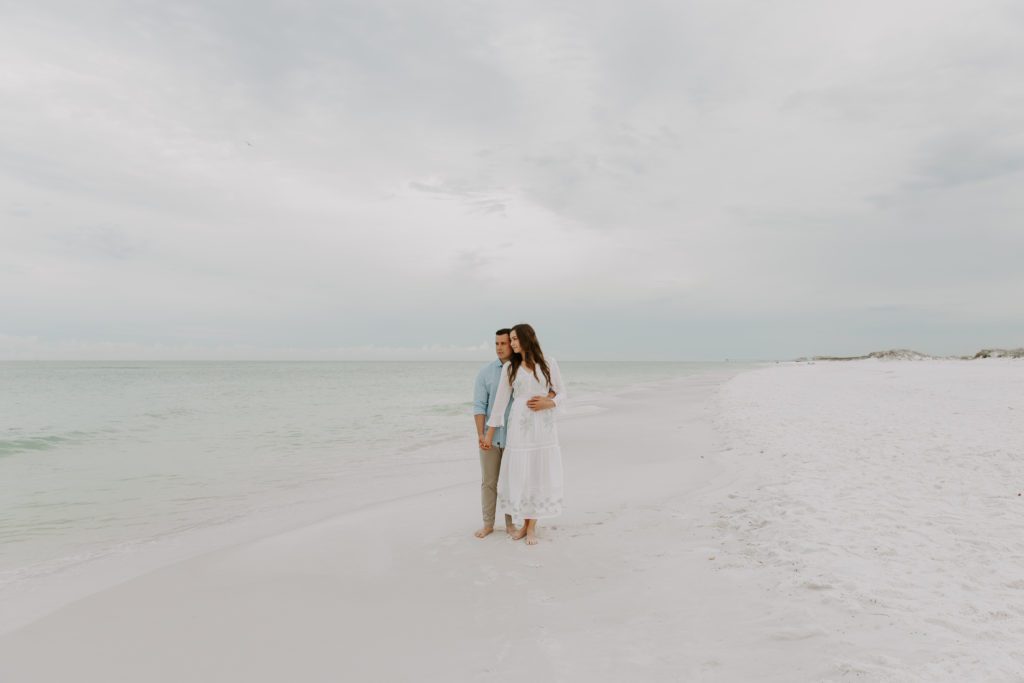 A couple standing on the empty beach leaning into each other and looking off into the distance taking in their surroundings during their Fort Walton Beach engagement photos