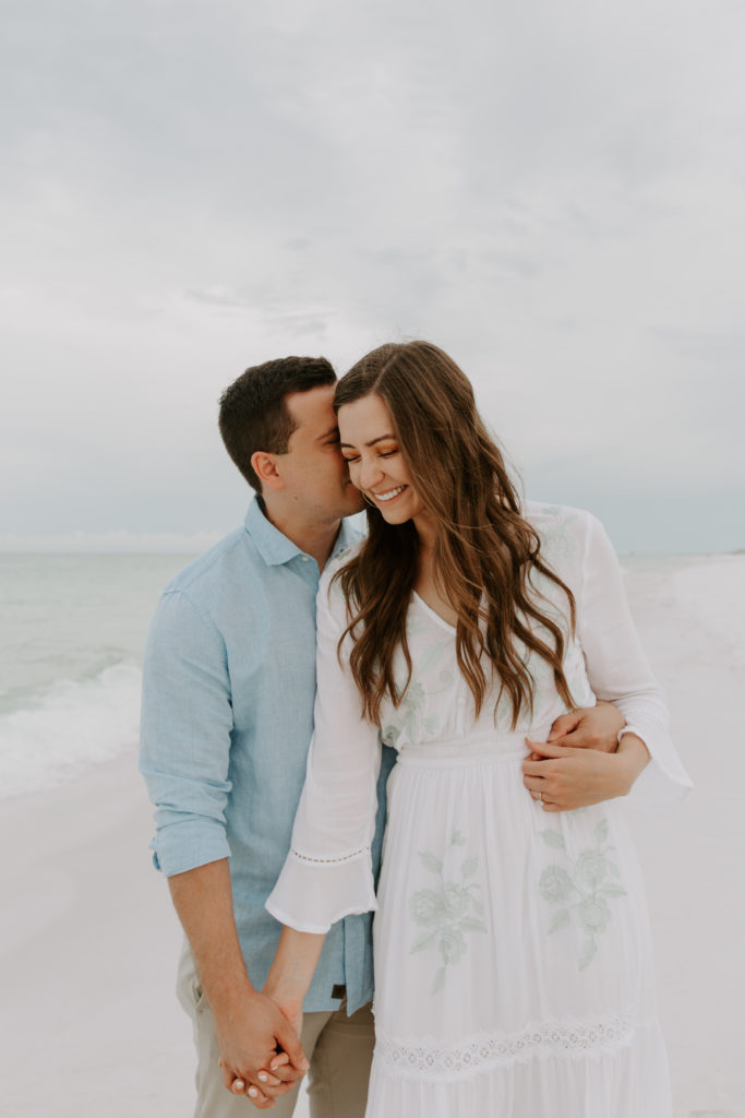 A newly engaged couple holding hands as the man whispers something in to his partners ear with the gulf of mexico in the background during their early beach photoshoot
