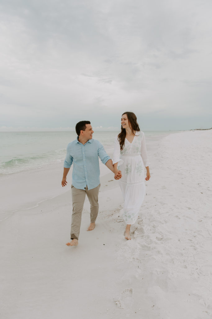 A couple holding hands and walking along the edge of the gulf of mexico looking at each other laughing