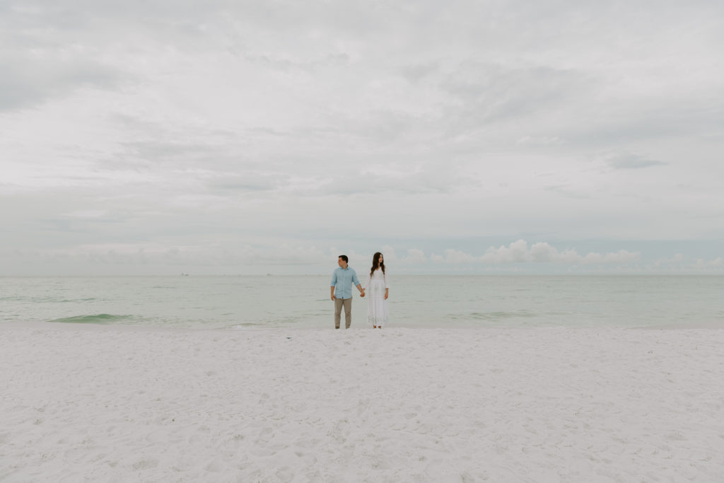 A couple standing at the waters edge in Florida holding hands and looking opposite directions of each other during their morning beach engagement photos