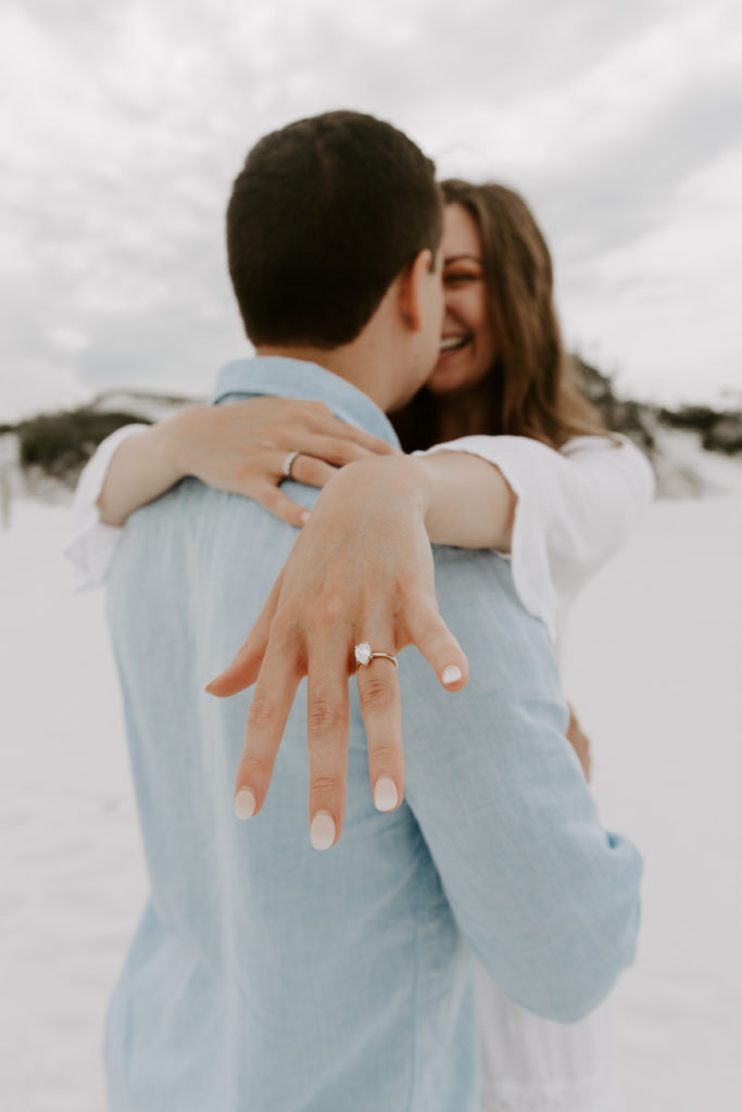 A woman holding her hand over her new fiance's shoulder showing off her engagement ring as they look at each other smiling