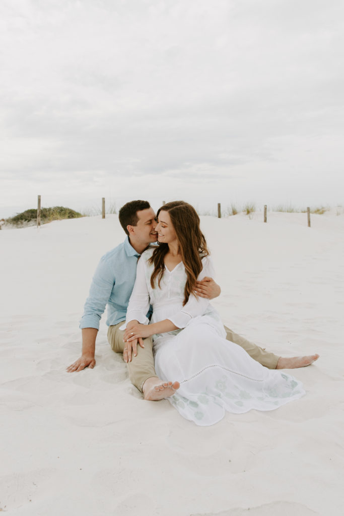 A couple sitting in front of beach sand dunes as he gives his partner a kiss on the cheek and she accepts it during their Florida engagement photos