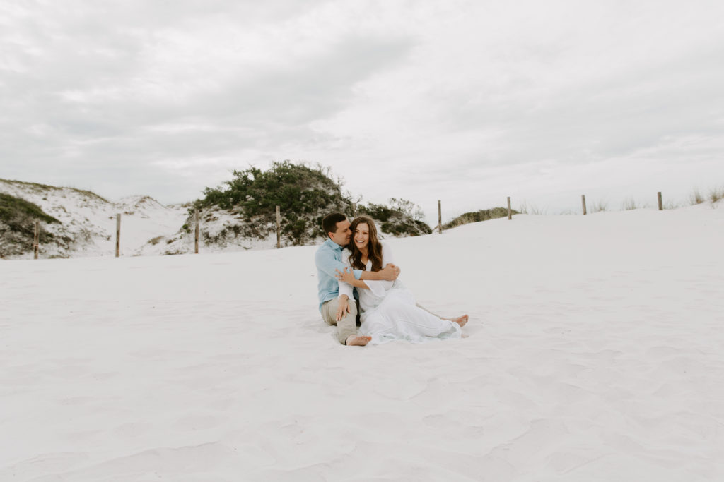 A couple sitting in the sand in front of large sand dunes with the man hugging onto his partner and her hugging him back during their beach proposal